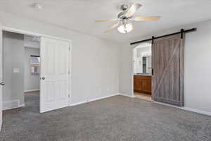 Unfurnished bedroom featuring ensuite bath, a barn door, ceiling fan, and carpet