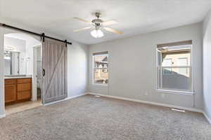 Unfurnished bedroom featuring a barn door, light colored carpet, ceiling fan, and ensuite bath