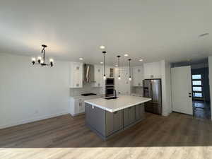 Kitchen with white cabinetry, hanging light fixtures, stainless steel appliances, a kitchen island with sink, and wall chimney range hood