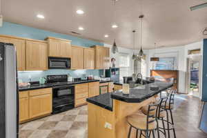 Kitchen featuring a breakfast bar, light brown cabinetry, a center island with sink, pendant lighting, and black appliances