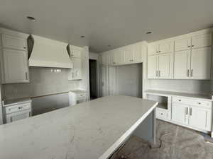 Kitchen with custom range hood, light stone countertops, white cabinetry, and backsplash