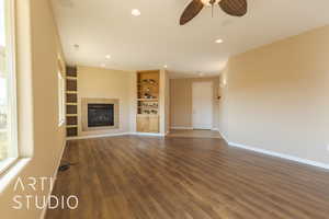 Unfurnished living room with built in shelves, dark lvp style floors, and a tiled fireplace and ceiling fan