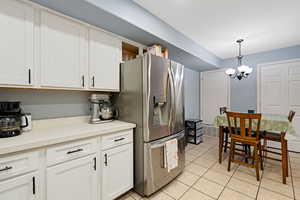 Kitchen with light tile patterned floors, stainless steel fridge, hanging light fixtures, a notable chandelier, and white cabinets