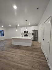 Kitchen featuring appliances with stainless steel finishes, decorative light fixtures, an island with sink, white cabinets, and dark wood-type flooring