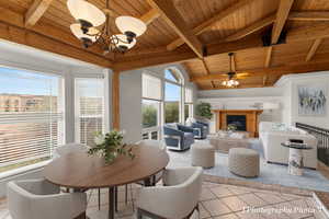 Dining area with ceiling fan with notable chandelier, vaulted ceiling with beams, light tile patterned floors, wood ceiling, and a brick fireplace