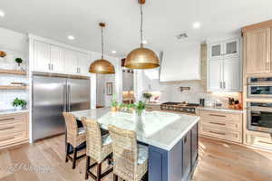 Kitchen with white & natural cabinetry, hanging light fixtures, stainless steel appliances, custom range hood, and a center island (black) with sink