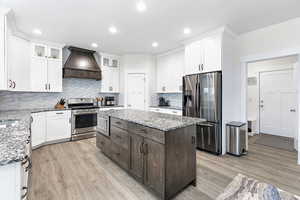 Kitchen with appliances with stainless steel finishes, white cabinetry, light stone counters, a kitchen island, and custom exhaust hood