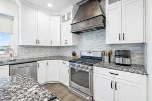 Kitchen featuring custom exhaust hood, dark stone countertops, white cabinets, and stainless steel appliances