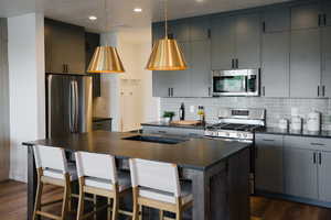 Kitchen featuring dark wood-type flooring, appliances with stainless steel finishes, gray cabinets, a kitchen island with sink, and decorative backsplash