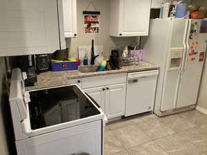 Kitchen with white cabinetry, sink, light tile patterned floors, light stone counters, and white appliances