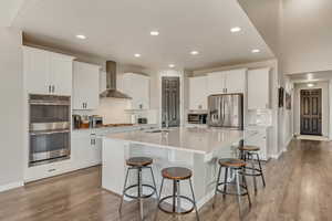 Kitchen featuring wall chimney range hood, an island with sink, white cabinets, and appliances with stainless steel finishes