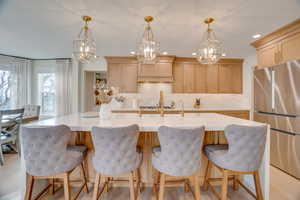 Kitchen with stainless steel fridge, decorative light fixtures, an island with sink, and light brown cabinets