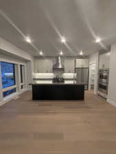 Kitchen featuring appliances with stainless steel finishes, wall chimney range hood, an island with sink, and white cabinets