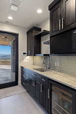 Butlers pantry off kitchen, featuring sink, backsplash, light stone counters, tile floors, and beverage cooler.