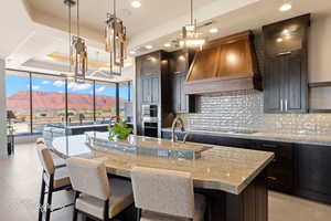 Kitchen featuring dark brown cabinetry, custom exhaust hood, a center island with sink, a mountain view, and a raised ceiling