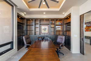 Home office with a raised ceiling, ceiling fan, and built in shelves