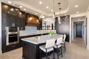 Kitchen featuring sink, light stone counters, a tray ceiling, custom range hood, and an island with sink