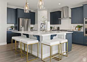 Kitchen with pendant lighting, wall chimney range hood, a breakfast bar area, appliances with stainless steel finishes, and an island with sink