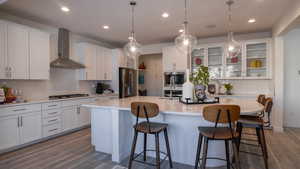 Kitchen with stainless steel refrigerator with ice dispenser, an island with sink, wall chimney range hood, black gas stovetop, and white cabinets