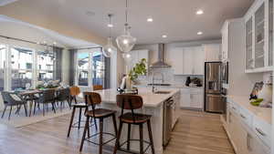 Kitchen featuring white cabinetry, wall chimney exhaust hood, hanging light fixtures, and a center island with sink