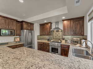 Kitchen featuring wall chimney exhaust hood, stainless steel appliances, a sink, recessed lighting, and light stone counters