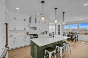 Kitchen with wall chimney range hood, a kitchen island with sink, hanging light fixtures, white cabinetry, and white fridge with ice dispenser