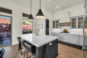 Kitchen with sink, light hardwood / wood-style floors, a center island with sink, decorative light fixtures, and custom exhaust hood