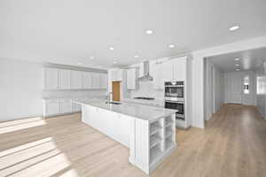 Kitchen with white cabinets, stainless steel double oven, a kitchen island with sink, and wall chimney range hood