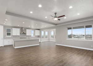 Kitchen with an island with sink, dark wood-type flooring, wall chimney range hood, and white cabinets