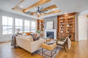 Living room with ceiling fan, a large fireplace, built in shelves, beamed ceiling, and light wood-type flooring