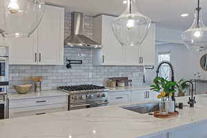 Kitchen featuring wall chimney range hood, stainless steel appliances, hanging light fixtures, and white cabinets