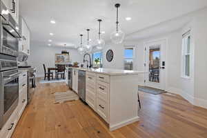 Kitchen featuring appliances with stainless steel finishes, decorative light fixtures, white cabinetry, light hardwood / wood-style floors, and a center island with sink