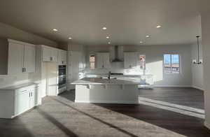 Kitchen with pendant lighting, wall chimney range hood, hardwood / wood-style flooring, an island with sink, and white cabinets