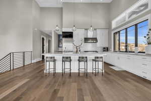 Kitchen with wall chimney range hood, a breakfast bar, an island with sink, white cabinets, and decorative light fixtures