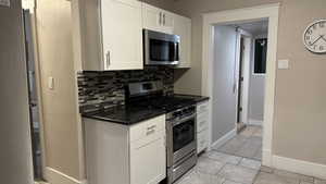 Kitchen featuring dark stone countertops, light tile patterned floors, white cabinets, stainless steel appliances, and backsplash