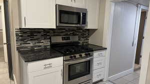 Kitchen with light tile patterned flooring, dark stone counters, white cabinets, stainless steel appliances, and backsplash