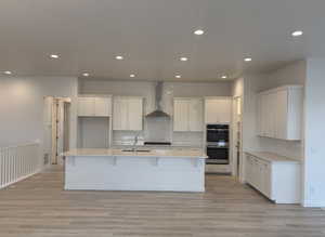 Kitchen featuring white cabinetry, wall chimney exhaust hood, a kitchen island with sink, and double oven