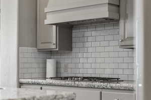 Kitchen with custom range hood, tasteful backsplash, stainless steel gas cooktop, and light stone countertops
