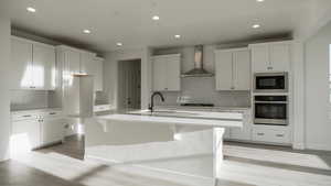 Kitchen with black microwave, stainless steel oven, an island with sink, white cabinets, and wall chimney exhaust hood