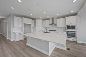 Kitchen featuring a spacious island, sink, wall chimney range hood, oven, and white cabinets
