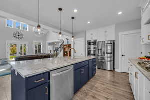 Kitchen featuring a kitchen island with sink, pendant lighting, wall ovens, white and blue cabinets, and appliances with stainless steel finishes