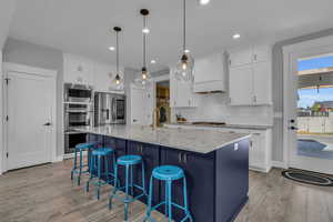 Kitchen featuring a kitchen island with sink, pendant lighting, wall ovens, white and blue cabinets, and appliances with stainless steel finishes