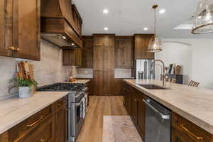 Kitchen with stainless steel appliances, sink, light wood-type flooring, premium range hood, and hanging light fixtures