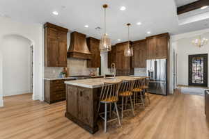 Kitchen featuring a center island with sink, appliances with stainless steel finishes, light hardwood / wood-style floors, premium range hood, and decorative light fixtures