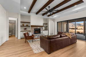 Living room featuring a stone fireplace, beamed ceiling, ceiling fan, light hardwood / wood-style flooring, and built in features