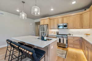 Kitchen featuring high quality appliances, light brown cabinetry, an island with sink, and hanging light fixtures