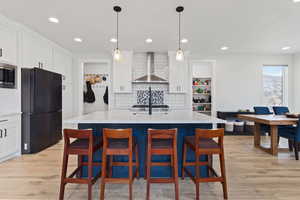 Kitchen featuring wall chimney exhaust hood, black fridge, light stone counters, decorative light fixtures, and a center island with sink