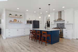 Kitchen featuring pendant lighting, white cabinets, a kitchen island with sink, stainless steel appliances, and wall chimney range hood