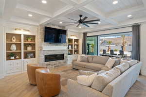 Living room with built in shelves, a stone fireplace, beamed ceiling, ceiling fan, and light parquet floors