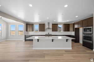 Kitchen featuring built in microwave, sink, stainless steel oven, a center island with sink, and wall chimney exhaust hood